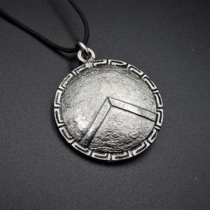 Könnte beinhalten: Eine silberne Halskette mit einem Anhänger in Form eines runden Schildes mit einem griechischen Schlüsselrand und einem erhabenen, strukturierten Design. Der Schild hat ein markantes V-förmiges Muster in der Mitte.