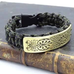 Puede incluir: Una pulsera de supervivencia con hebilla de pl&aacute;stico negro y cord&oacute;n trenzado negro y verde oliva. La pulsera presenta una placa met&aacute;lica dorada con un dise&ntilde;o floral grabado. La pulsera est&aacute; sobre un libro antiguo.