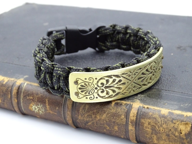 Puede incluir: Una pulsera de supervivencia con hebilla de pl&aacute;stico negro y cord&oacute;n trenzado negro y verde oliva. La pulsera presenta una placa met&aacute;lica dorada con un dise&ntilde;o floral grabado. La pulsera est&aacute; sobre un libro antiguo.