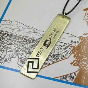 May include: A gold-coloured metal pendant necklace with a rectangular tag featuring an engraved image of a ship and the text "&Epsilon;&Sigma;&Sigma;&Epsilon;&Tau;' &Eta;&Mu;&Delta;&Alpha;&Rho; &Omicron;&Mu;&Eta;&Rho;&Omicron;&Sigma;".