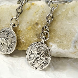 May include: Two silver keychains with coin-shaped charms. One charm features a profile of a man with a laurel wreath, the other charm features a woman riding a horse. Both charms have text around the edges.