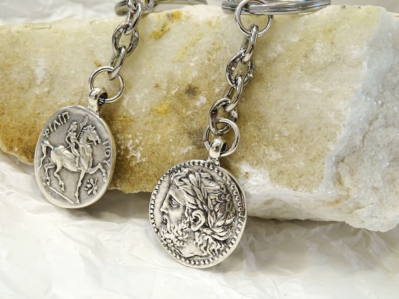 May include: Two silver keychains with coin-shaped charms. One charm features a profile of a man with a laurel wreath, the other charm features a woman riding a horse. Both charms have text around the edges.