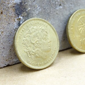 Puede incluir: Dos monedas de color dorado con escritura griega. La moneda de la izquierda presenta un perfil de un hombre con una corona y el texto "ME&Gamma;A&Sigma; A&Lambda;E&Xi;AN&Delta;PO&Sigma; &Beta;&Alpha;&Sigma;&Iota;&Lambda;&Epsilon;&Upsilon;&Sigma; &Mu;&Alpha;&Kappa;&Epsilon;&Delta;&Omicron;&Nu;&Omega;&Nu;". La moneda de la derecha presenta un sol con rayos y el texto "100".