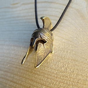 May include: A gold-colored metal pendant in the shape of a Spartan helmet. The helmet has a textured surface and a pointed crest. The pendant is hanging from a black cord.