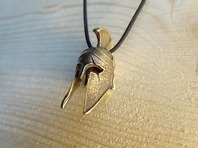 May include: A gold-colored metal pendant in the shape of a Spartan helmet. The helmet has a textured surface and a pointed crest. The pendant is hanging from a black cord.