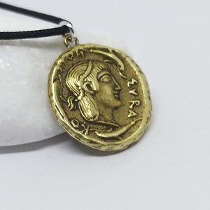 Puede incluir: Un collar con un colgante en forma de moneda griega antigua de color dorado, que representa el perfil de una mujer y la inscripci&oacute;n "&Sigma;&Upsilon;&Rho;&Alpha;" en el borde.