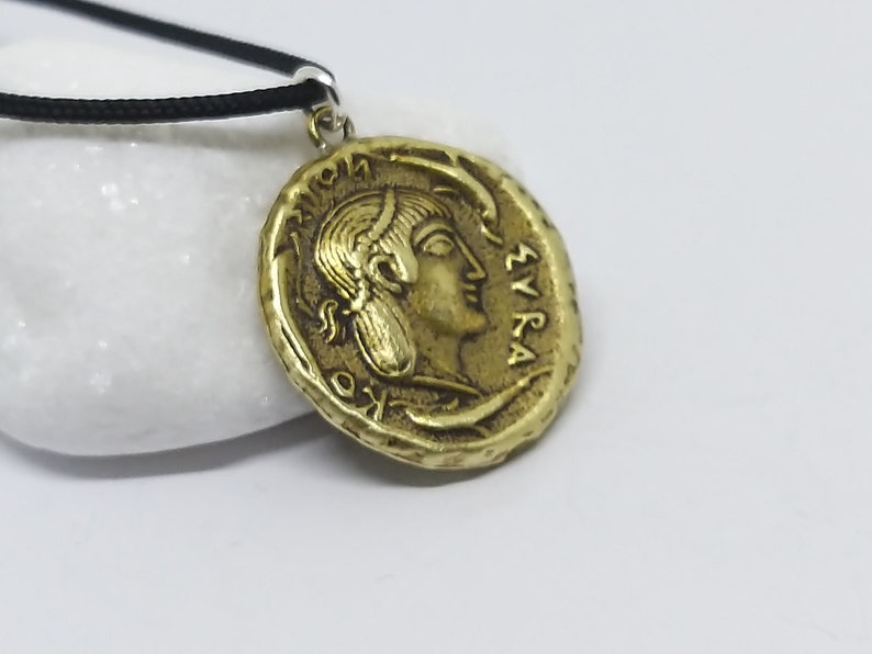 Puede incluir: Un collar con un colgante en forma de moneda griega antigua de color dorado, que representa el perfil de una mujer y la inscripci&oacute;n "&Sigma;&Upsilon;&Rho;&Alpha;" en el borde.