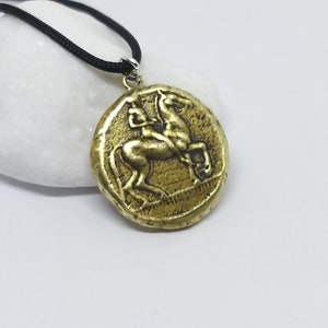 Puede incluir: Un collar con un colgante redondo en forma de moneda que presenta una imagen en relieve de un caballo. El colgante est&aacute; en un cord&oacute;n negro.