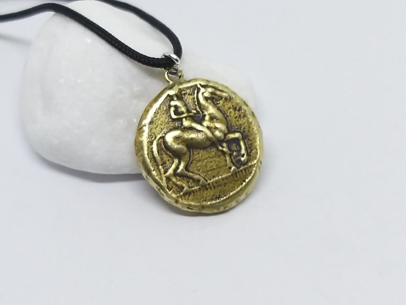 Puede incluir: Un collar con un colgante redondo en forma de moneda que presenta una imagen en relieve de un caballo. El colgante est&aacute; en un cord&oacute;n negro.