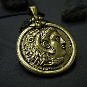 Op de afbeelding: Een gouden ketting met een ronde muntvormige hanger met een gedetailleerd profiel van een gezicht. De ketting is aan een zwart koord.