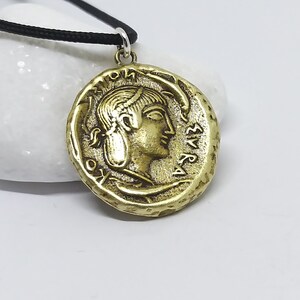 Puede incluir: Collar con colgante redondo en tono dorado, que representa una moneda con el perfil de una mujer y letras griegas. El collar est&aacute; en un cord&oacute;n negro.