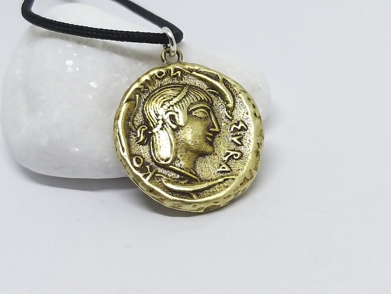 Puede incluir: Collar con colgante redondo en tono dorado, que representa una moneda con el perfil de una mujer y letras griegas. El collar est&aacute; en un cord&oacute;n negro.