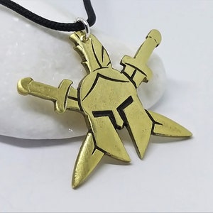 May include: A gold-toned pendant necklace featuring a Spartan helmet design with two crossed swords. The necklace is on a black cord.