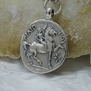 May include: Silver pendant featuring a woman riding a horse, with the inscription "AININ TOY" around the edge.