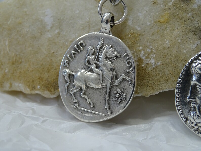 May include: Silver pendant featuring a woman riding a horse, with the inscription "AININ TOY" around the edge.