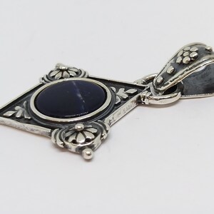 May include: A silver pendant with a dark blue gemstone set in a diamond-shaped frame with floral details. The pendant has a silver bail with a floral design.