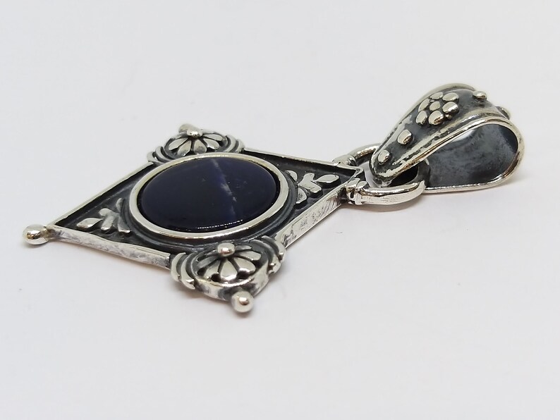 May include: A silver pendant with a dark blue gemstone set in a diamond-shaped frame with floral details. The pendant has a silver bail with a floral design.
