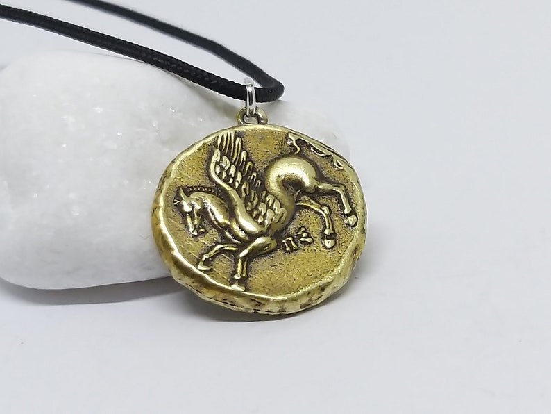 K&ouml;nnte beinhalten: Eine goldfarbene Halskette mit einem Anh&auml;nger in Form eines gefl&uuml;gelten Pferdes, oder Pegasus. Der Anh&auml;nger ist rund und hat eine strukturierte Oberfl&auml;che. Die Halskette ist an einer schwarzen Schnur.