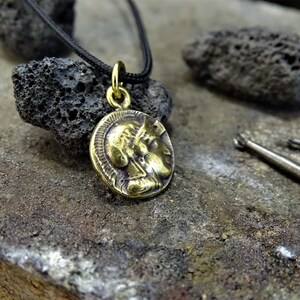 Puede incluir: Un collar con un colgante redondo en forma de moneda que presenta un perfil de cabeza. El collar est&aacute; en un cord&oacute;n negro y est&aacute; descansando sobre una superficie gris y rugosa.