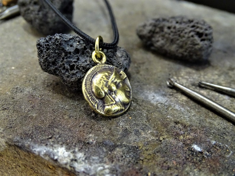 Puede incluir: Un collar con un colgante redondo en forma de moneda que presenta un perfil de cabeza. El collar est&aacute; en un cord&oacute;n negro y est&aacute; descansando sobre una superficie gris y rugosa.