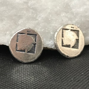 Puede incluir: Dos pendientes redondos plateados. Cada pendiente presenta un dise&ntilde;o cuadrado con una imagen en relieve en el interior. Los pendientes tienen una superficie texturizada y ligeramente irregular. El n&uacute;mero "925" est&aacute; estampado en la parte superior de cada pendiente.