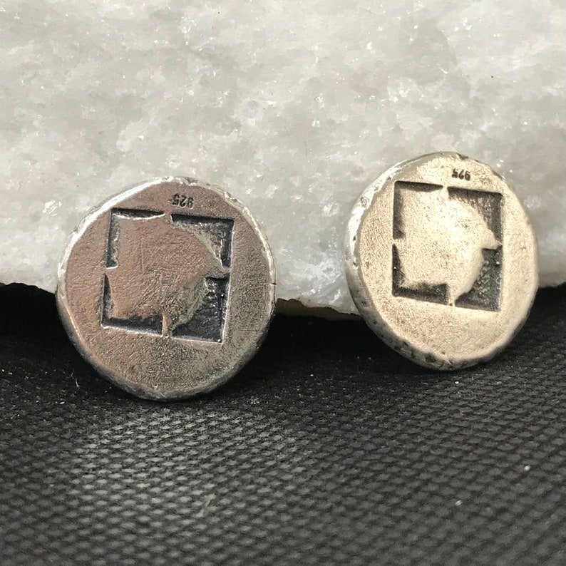 Puede incluir: Dos pendientes redondos plateados. Cada pendiente presenta un dise&ntilde;o cuadrado con una imagen en relieve en el interior. Los pendientes tienen una superficie texturizada y ligeramente irregular. El n&uacute;mero "925" est&aacute; estampado en la parte superior de cada pendiente.