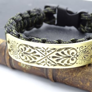 Puede incluir: Una pulsera de supervivencia con un cord&oacute;n trenzado negro y verde oliva. La pulsera presenta una placa decorativa de metal dorado con un dise&ntilde;o floral grabado. El cierre es negro.
