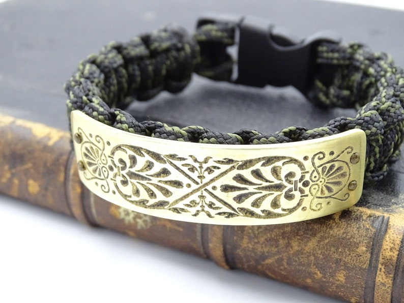 Puede incluir: Una pulsera de supervivencia con un cord&oacute;n trenzado negro y verde oliva. La pulsera presenta una placa decorativa de metal dorado con un dise&ntilde;o floral grabado. El cierre es negro.