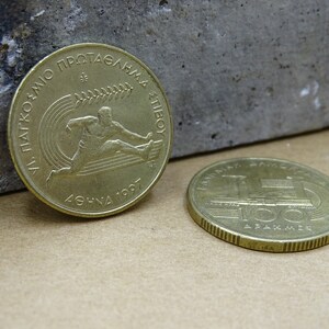 May include: Two gold-colored Greek coins. The first coin features a runner and the text "XI PANATHENAIKO PROTAGONMA ATHINON 1997". The second coin features a train and the text "&Epsilon;&Lambda;&Lambda;&Eta;&Nu;&Iota;&Kappa;&Eta; &Delta;&Eta;&Mu;&Omicron;&Kappa;&Rho;&Alpha;&Tau;&Iota;&Alpha; 100 &Delta;&Rho;&Alpha;&Chi;&Mu;&Epsilon;&Sigma;".