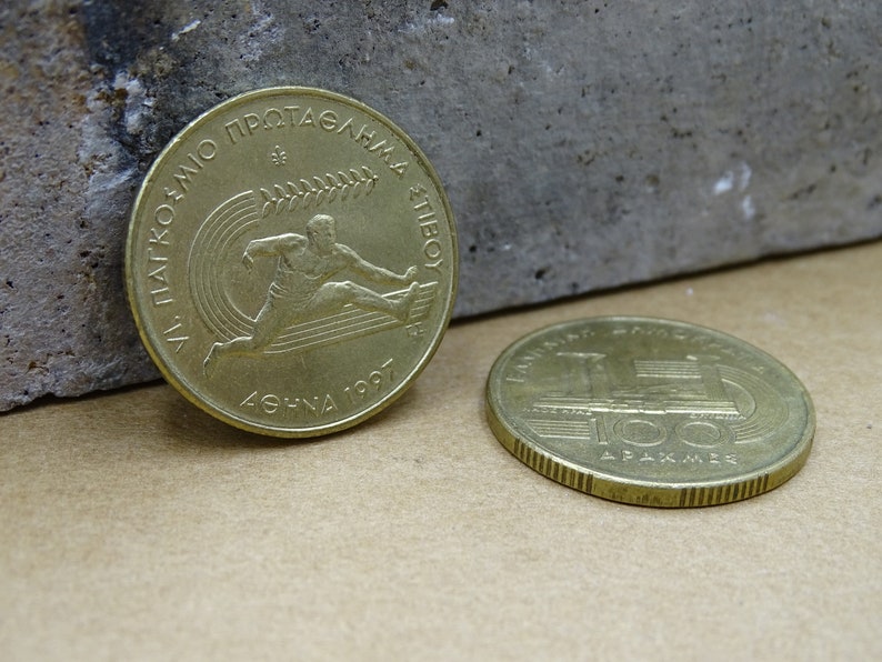 May include: Two gold-colored Greek coins. The first coin features a runner and the text "XI PANATHENAIKO PROTAGONMA ATHINON 1997". The second coin features a train and the text "&Epsilon;&Lambda;&Lambda;&Eta;&Nu;&Iota;&Kappa;&Eta; &Delta;&Eta;&Mu;&Omicron;&Kappa;&Rho;&Alpha;&Tau;&Iota;&Alpha; 100 &Delta;&Rho;&Alpha;&Chi;&Mu;&Epsilon;&Sigma;".