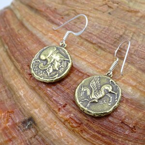May include: A pair of gold-toned earrings featuring a winged horse and a mythical creature on round coin-shaped charms. The earrings are suspended from silver-toned hooks.