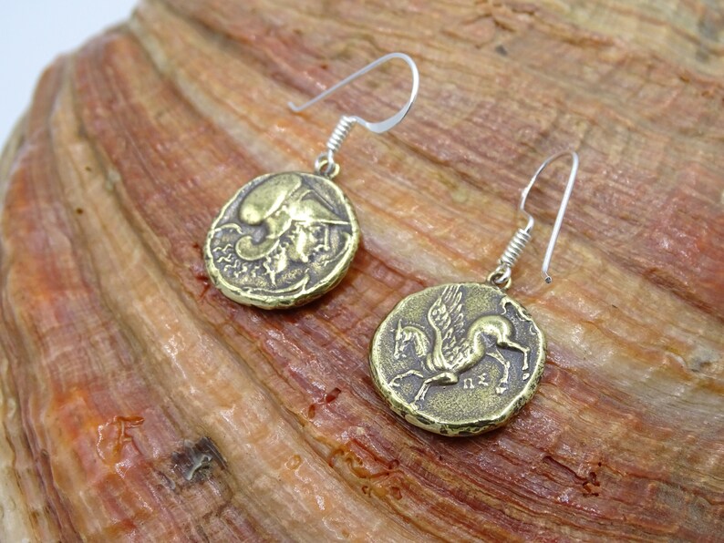 May include: A pair of gold-toned earrings featuring a winged horse and a mythical creature on round coin-shaped charms. The earrings are suspended from silver-toned hooks.