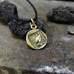 Puede incluir: Un collar con un colgante redondo en forma de moneda con un dise&ntilde;o de p&aacute;jaro estilizado. El collar est&aacute; en un cord&oacute;n negro.