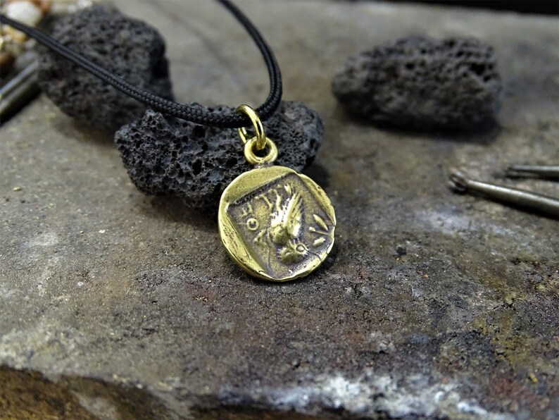 Puede incluir: Un collar con un colgante redondo en forma de moneda con un dise&ntilde;o de p&aacute;jaro estilizado. El collar est&aacute; en un cord&oacute;n negro.