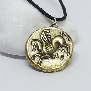 K&ouml;nnte beinhalten: Eine goldfarbene Halskette mit einem Anh&auml;nger in Form eines gefl&uuml;gelten Pferdes, oder Pegasus. Der Anh&auml;nger ist rund und hat eine strukturierte Oberfl&auml;che. Die Halskette ist an einer schwarzen Schnur.