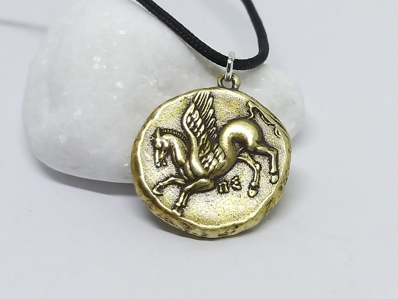 K&ouml;nnte beinhalten: Eine goldfarbene Halskette mit einem Anh&auml;nger in Form eines gefl&uuml;gelten Pferdes, oder Pegasus. Der Anh&auml;nger ist rund und hat eine strukturierte Oberfl&auml;che. Die Halskette ist an einer schwarzen Schnur.