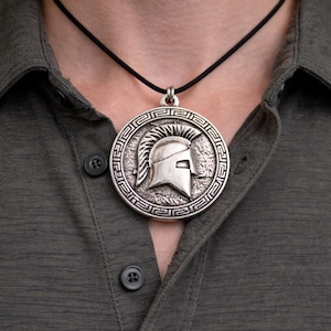 Puede incluir: Collar con colgante plateado con diseño de casco espartano. El colgante redondo cuelga de un cordón negro, usado sobre una camisa verde oscuro abotonada. El colgante presenta un diseño detallado en relieve.