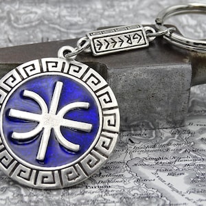 May include: A silver-colored keychain with a round pendant featuring a blue center and a white geometric design. The pendant is attached to a key ring and a rectangular piece with the word "GREECE" in Greek letters.