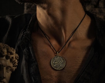 May include: A bronze-colored pendant necklace featuring a detailed profile of a classical figure. The pendant hangs from a black cord. The necklace is worn against a dark background, highlighting the intricate design of the pendant.