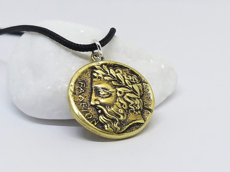 May include: A gold-toned pendant necklace with a round coin-shaped charm featuring a detailed profile of a man's face with a laurel wreath. The inscription "PANEION" is visible on the charm.