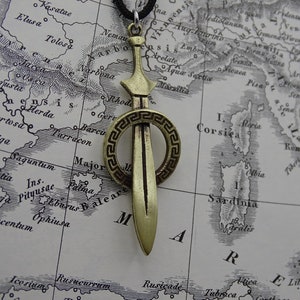 Może przedstawiać: Złoty naszyjnik z zawieszką w kształcie miecza, ozdobiony okrągłym, dekoracyjnym wzorem greckiego klucza. Zawieszka znajduje się na brązowej, zabytkowej mapie.