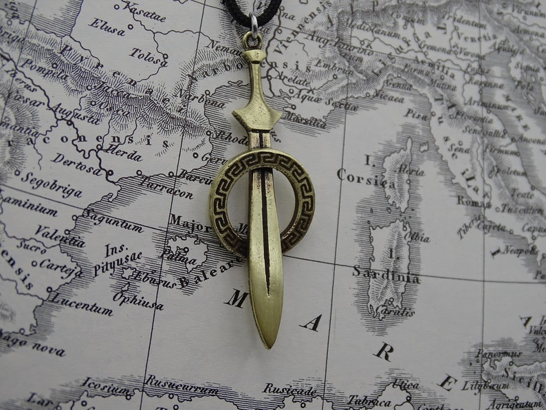 Może przedstawiać: Złoty naszyjnik z zawieszką w kształcie miecza, ozdobiony okrągłym, dekoracyjnym wzorem greckiego klucza. Zawieszka znajduje się na brązowej, zabytkowej mapie.