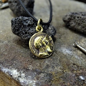 Puede incluir: Un collar con un colgante redondo en forma de moneda que presenta el perfil de la cara de una mujer. El collar est&aacute; en un cord&oacute;n negro y est&aacute; descansando sobre una superficie rugosa de color gris oscuro.