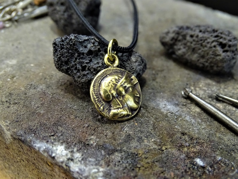 Puede incluir: Un collar con un colgante redondo en forma de moneda que presenta el perfil de la cara de una mujer. El collar est&aacute; en un cord&oacute;n negro y est&aacute; descansando sobre una superficie rugosa de color gris oscuro.