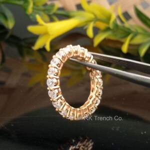 Peut inclure: Une bague en or avec une rangée de pierres rondes transparentes. La bague est tenue par une pince à épiler et le texte "RK Trench Co." est visible dans le coin inférieur droit.