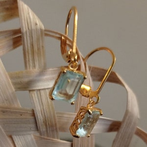 May include: Gold earrings with light blue gemstone drops. The earrings have a simple design with a small, rectangular gemstone hanging from a gold hook.