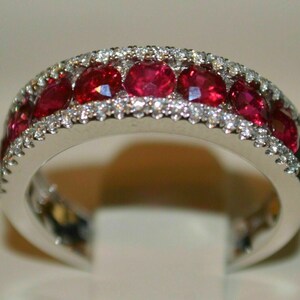 May include: A silver ring featuring a row of oval-cut, vibrant red gemstones, likely rubies, set between two rows of smaller, sparkling white gemstones, possibly diamonds. The ring has a polished, reflective surface.