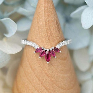 May include: A silver ring with a curved band and a cluster of five red marquise-shaped gemstones. The ring is set on a wooden display stand.