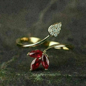 Könnte beinhalten: Goldfarbener Ring mit Blattmotiv. Ein Blatt ist mit kleinen, klaren Steinen besetzt, das andere mit drei tiefroten, marquiseförmigen Steinen. Der Ring verläuft elegant, wodurch ein von der Natur inspiriertes Design entsteht.