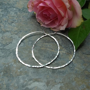 May include: Two silver hoop earrings with a hammered texture, displayed on a grey stone surface. A pink rose with green leaves is in the background. The earrings are a classic jewellery item, suitable for everyday wear or special occasions.